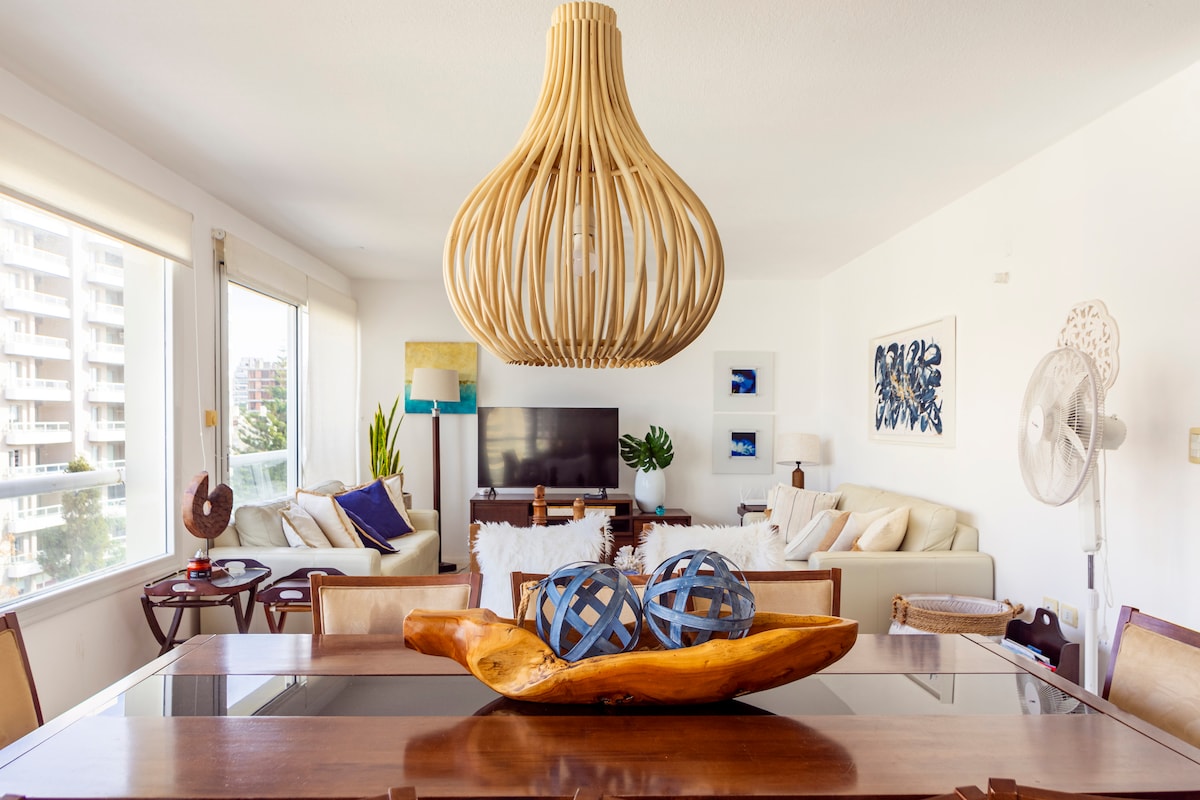 A bright and open living area features a wooden dining table centered beneath an artistic pendant light. Two comfortable seating areas are visible, with soft couches and decorative accents. Large windows allow natural light to illuminate the space, enhancing the room's airy feel.