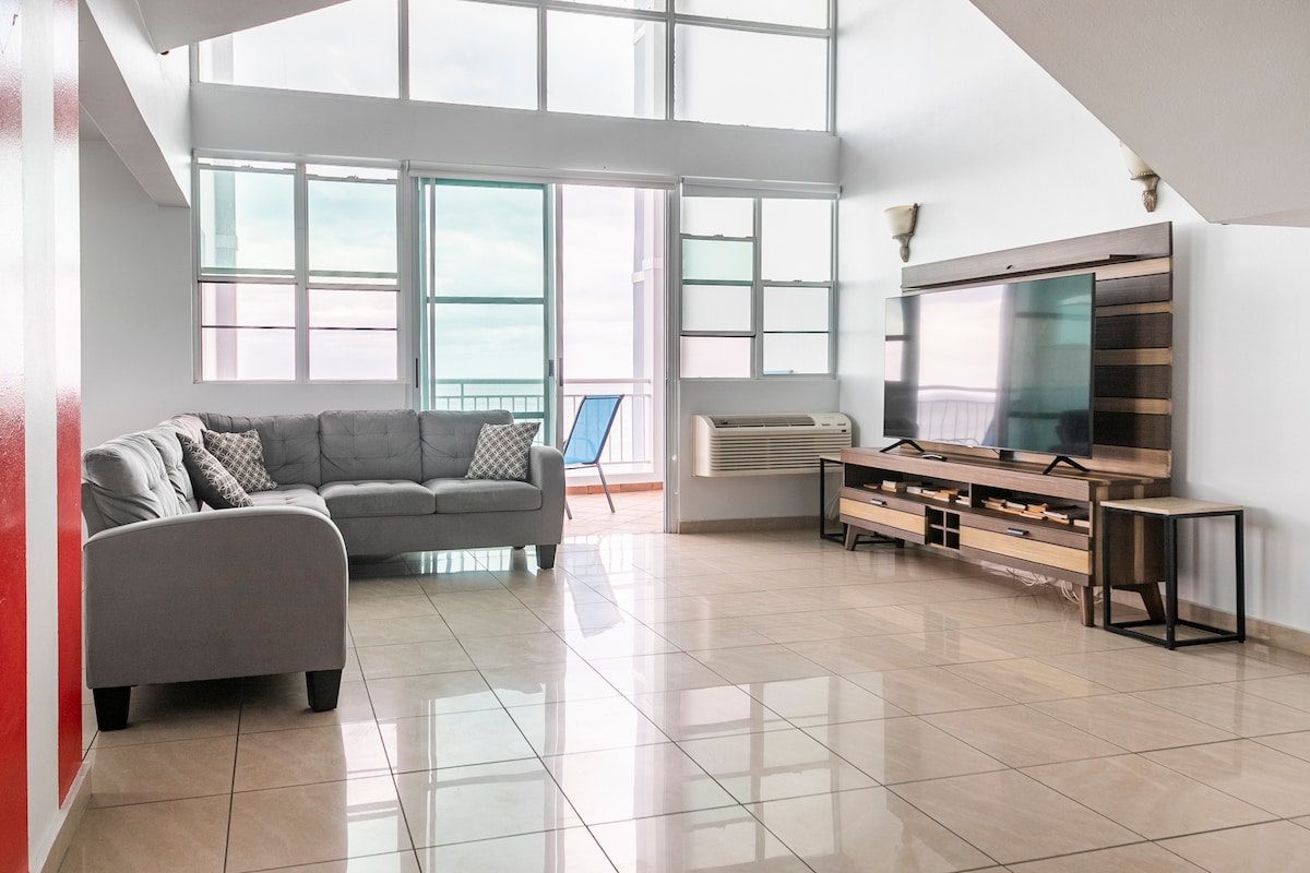 A spacious living area features large windows that allow natural light to fill the space, providing direct access to a balcony with bay views. A comfortable gray sofa is positioned facing a modern entertainment unit with a flat-screen TV, while neutral tile flooring reflects the light.