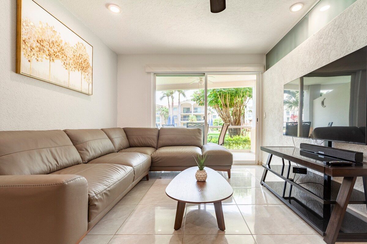 A spacious living area features a large sectional sofa in a neutral tone, complemented by a round coffee table. A flat-screen TV is mounted on the wall, and sliding glass doors lead out to a green outdoor space, inviting natural light into the room.