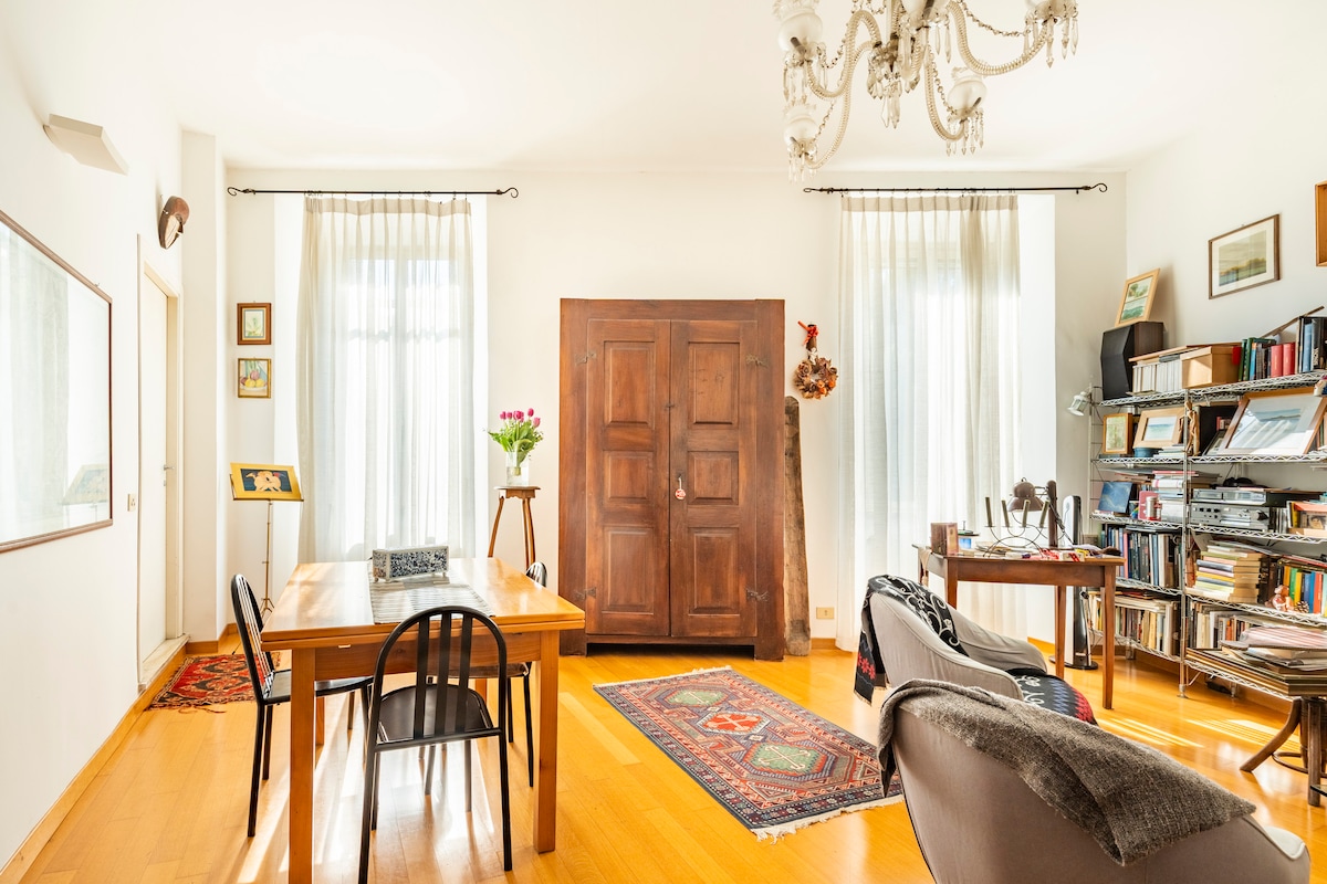 A spacious living area features a wooden dining table accompanied by chairs, positioned on a vibrant area rug. Soft natural light enters through large windows adorned with sheer curtains. A bookshelf is visible, filled with various books, adding character to the room.