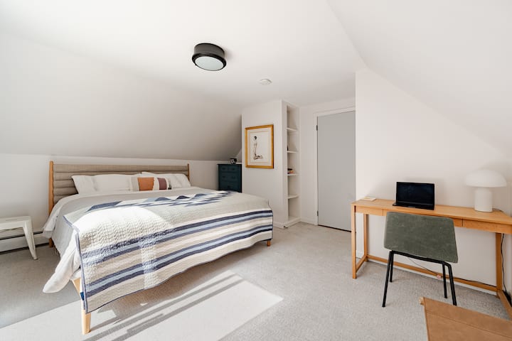 Bedroom 1 — Balcony View: King bedroom with desk, cozy bedding, blackout curtains, AC unit, and a small balcony overlooking the front of the property.