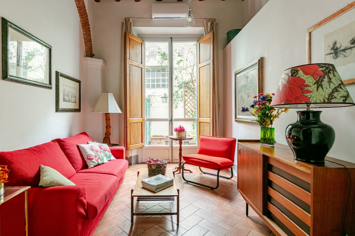 A bright living room displays a red sofa with decorative cushions, accompanied by a matching red chair. Natural light floods the space through large open windows, which lead to an outdoor area. Artwork and a lamp add character to the room, enhancing its welcoming atmosphere.