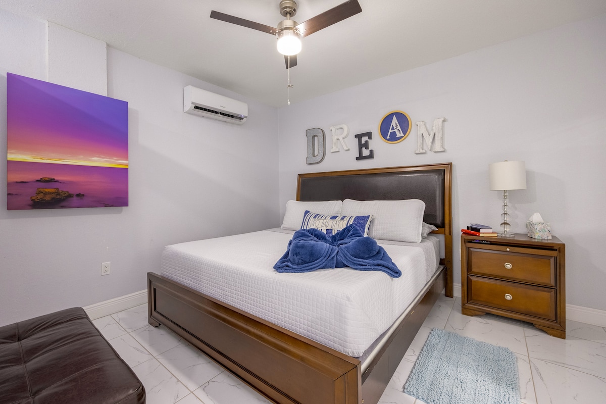 A well-appointed sleeping area features a spacious bed with a tasteful blue throw positioned at the foot. A piece of colorful wall art is displayed nearby, and a nightstand holds a lamp. A ceiling fan and air conditioning unit are visible, providing comfort.