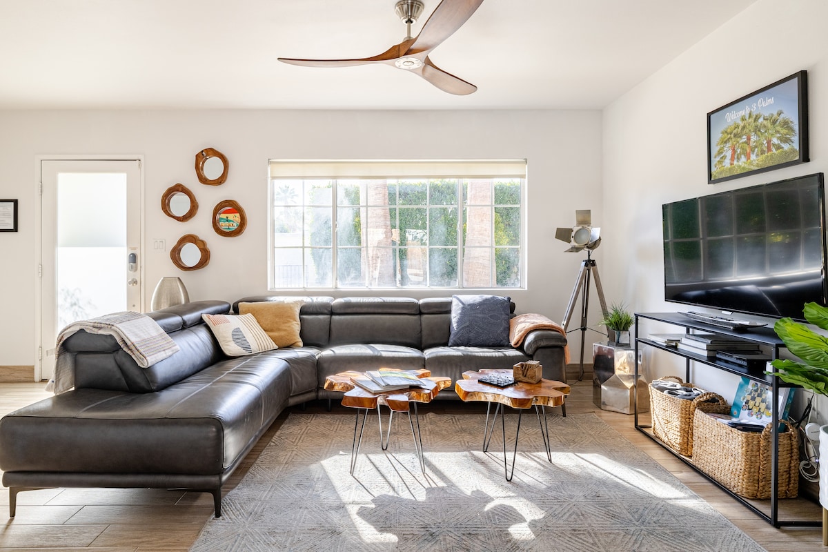 A spacious living area is highlighted by a large sectional sofa with neutral tones. A flat-screen TV is positioned on a media console, while decorative wooden tables provide a rustic touch. Natural light streams through the extensive windows, illuminating the room.