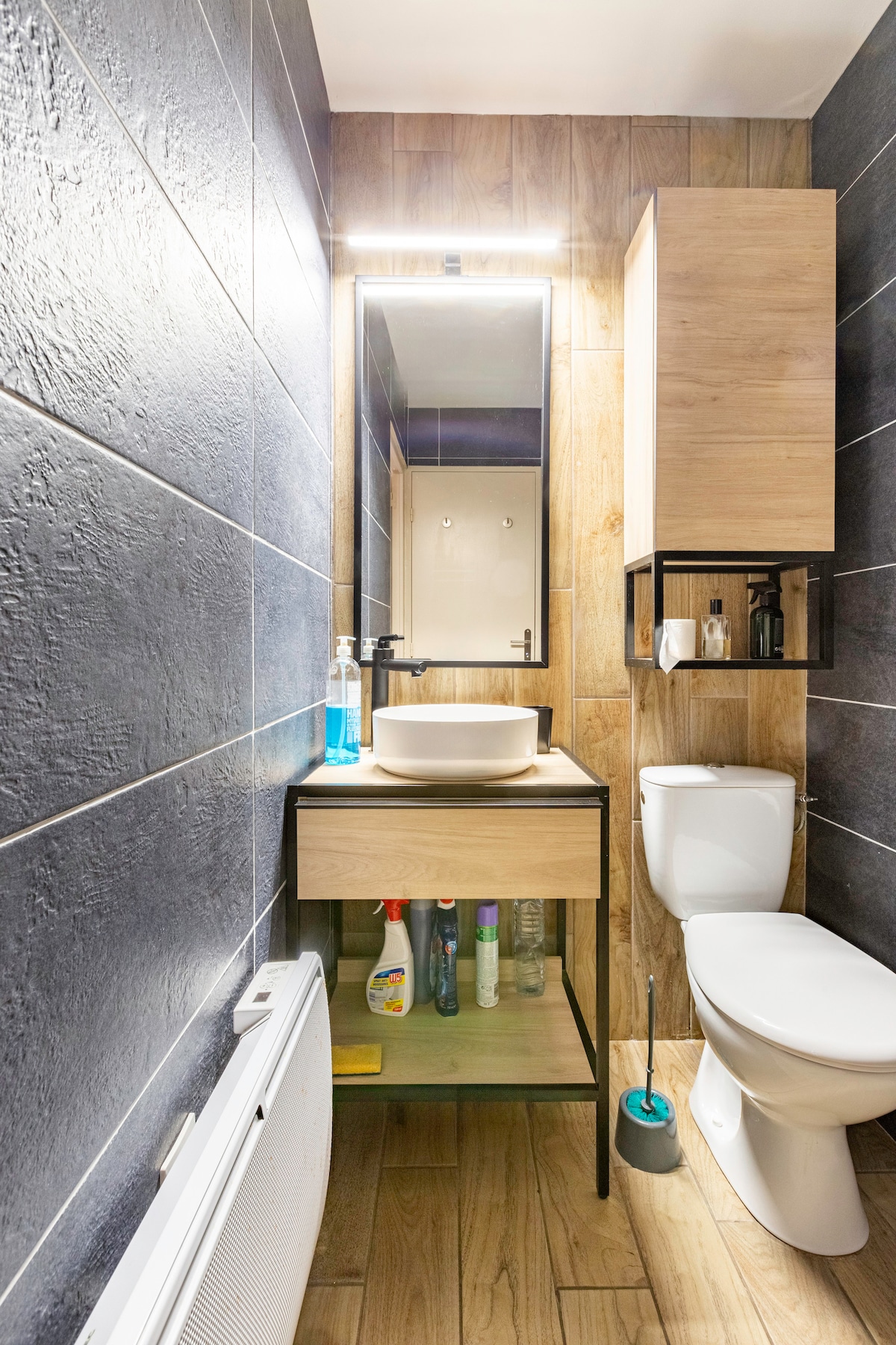 The bathroom features a modern design with dark tiled walls and a wooden vanity. A round sink sits atop the vanity, which holds various cleaning supplies. The toilet is positioned adjacent to the vanity, and a mirror is illuminated by overhead lighting.