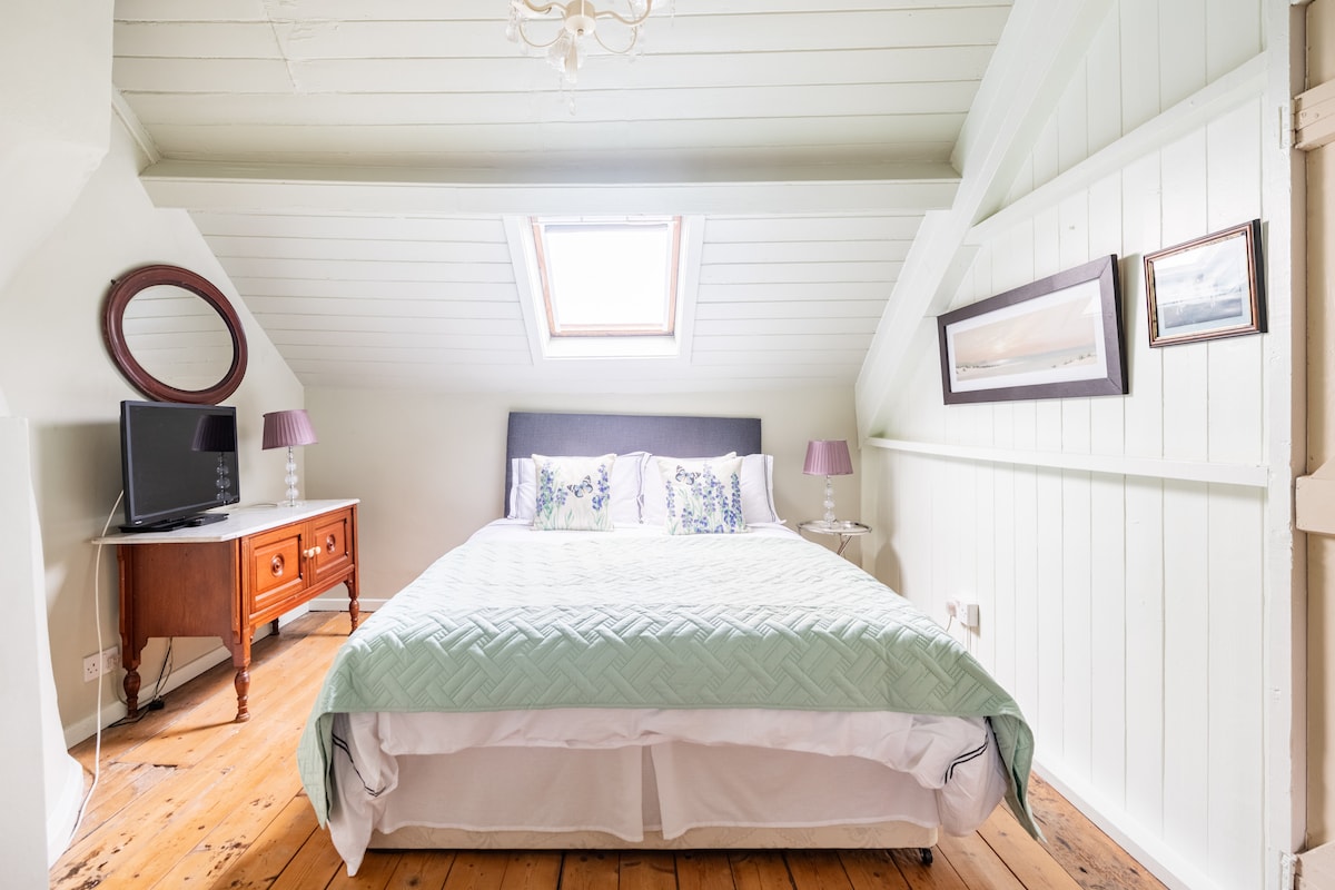 An upstairs bedroom is depicted, featuring a spacious bed with a light green bedspread. Natural light enters through a skylight, highlighting the wooden flooring. A TV is positioned on a wooden dresser, complemented by framed pictures on the walls.