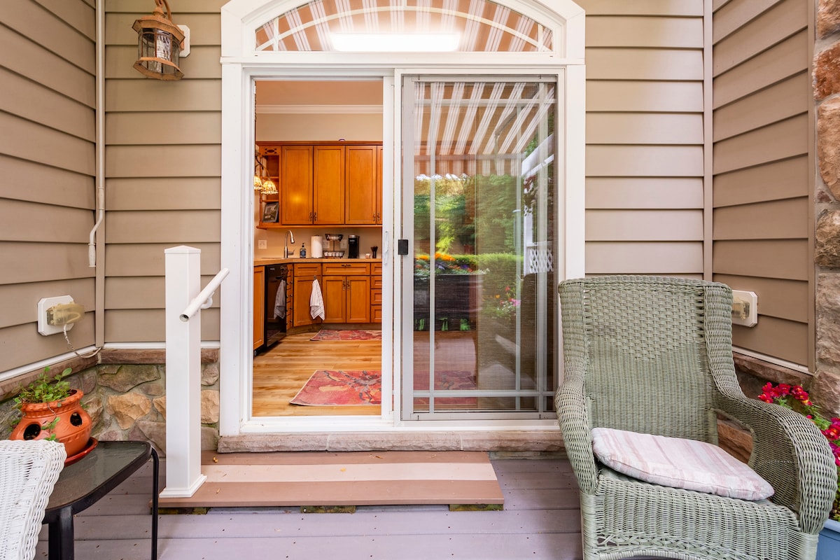 The entrance features sliding glass doors leading to the kitchen, framed by a patterned awning. A comfortable green wicker chair is positioned next to a small side table. Potted flowers add a splash of color to the inviting outdoor space.