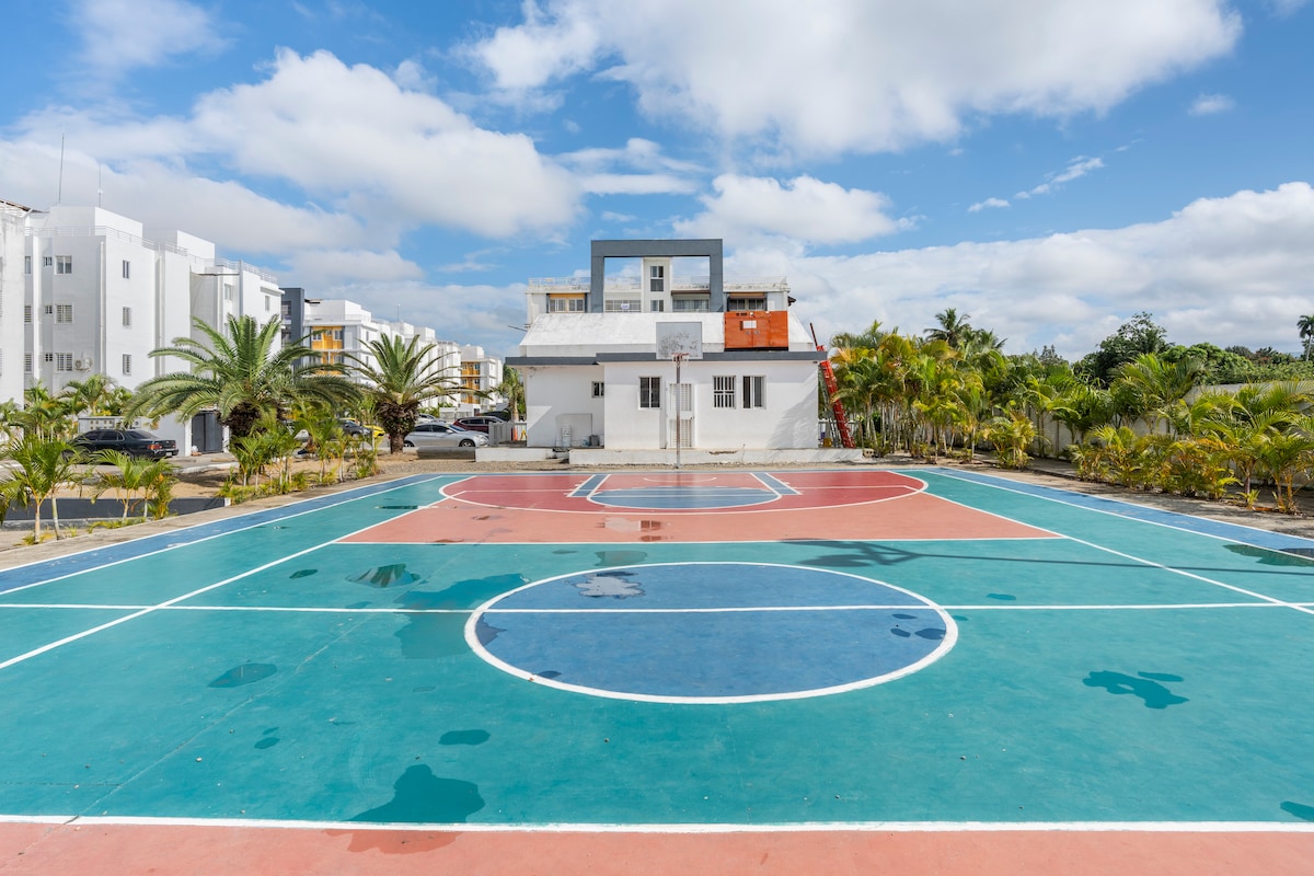 A basketball court is shown with vibrant red and blue sections marked by white lines. Surrounding palm trees enhance the setting, while a building stands in the background, providing context. The surface appears clean and well-maintained, suitable for recreational activities.