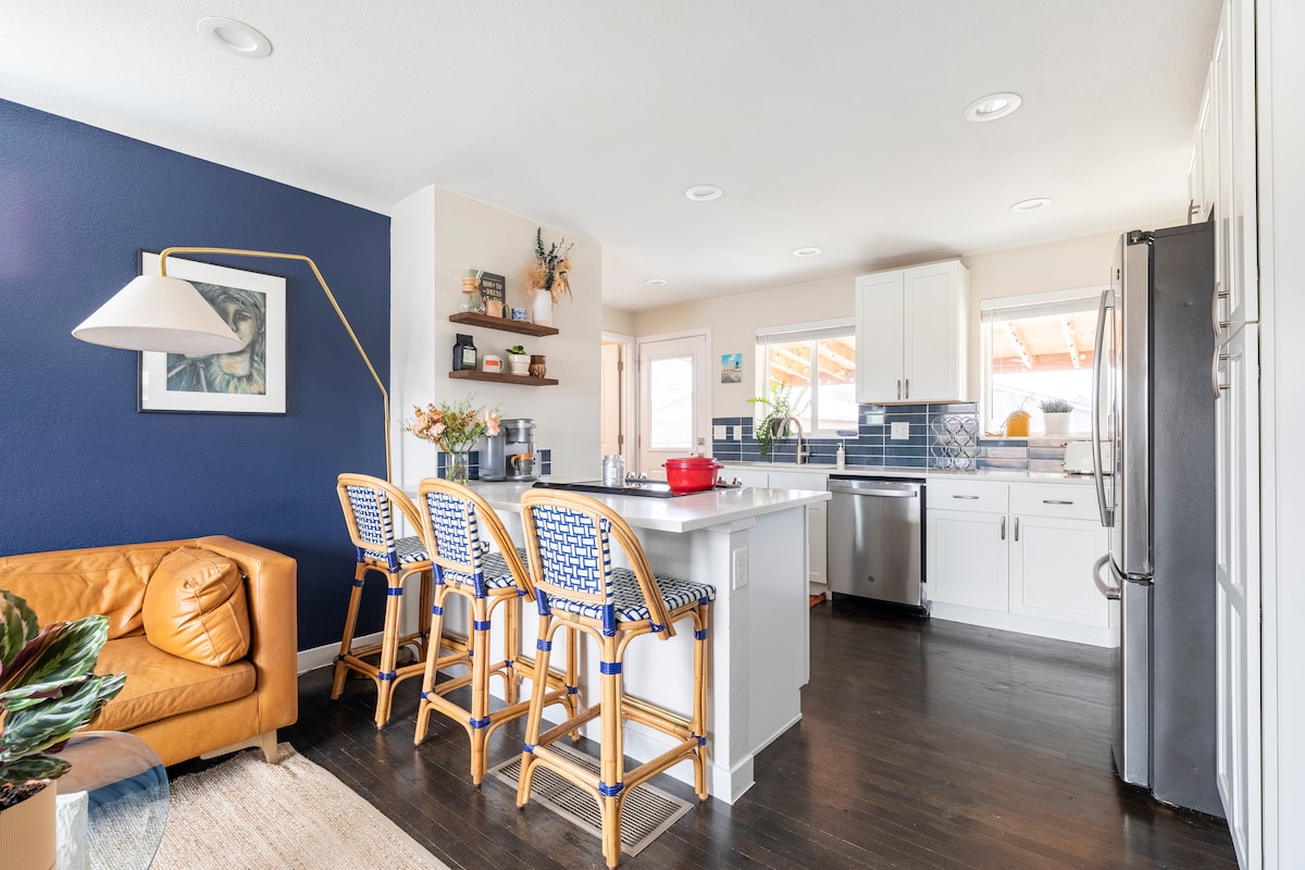 An open kitchen and dining area feature modern appliances against a navy accent wall. The kitchen island is complemented by four woven bar stools, and natural light fills the space through large windows. A cozy couch and decorative plants enhance the inviting atmosphere.