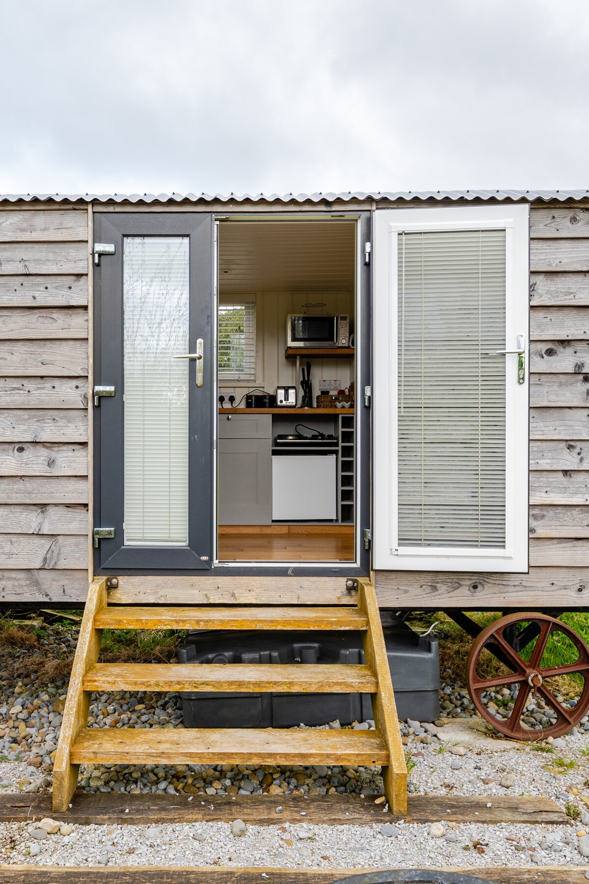 The entrance features a set of double doors that open onto a wooden staircase. The rustic exterior is complemented by gray and white panels. A glimpse of the kitchen area is visible inside, showcasing modern appliances and a welcoming layout.