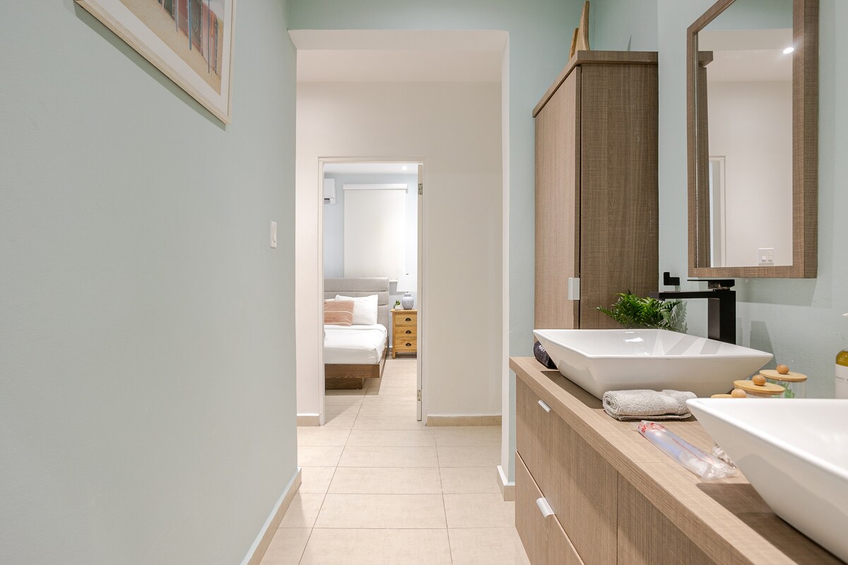 A spacious corridor with soft blue walls leads to the living area, featuring a wooden cabinet with a modern sink. Natural light illuminates the space, creating a calm atmosphere. A doorway in the background provides a glimpse of the bedroom.