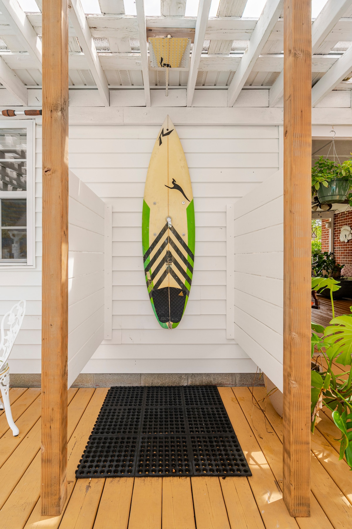 An outdoor shower area is presented, featuring a surfboard mounted on white walls. The space is framed by wooden posts, with a textured black mat laid below. The surrounding area includes light-colored wooden decking and hints of greenery from planters.