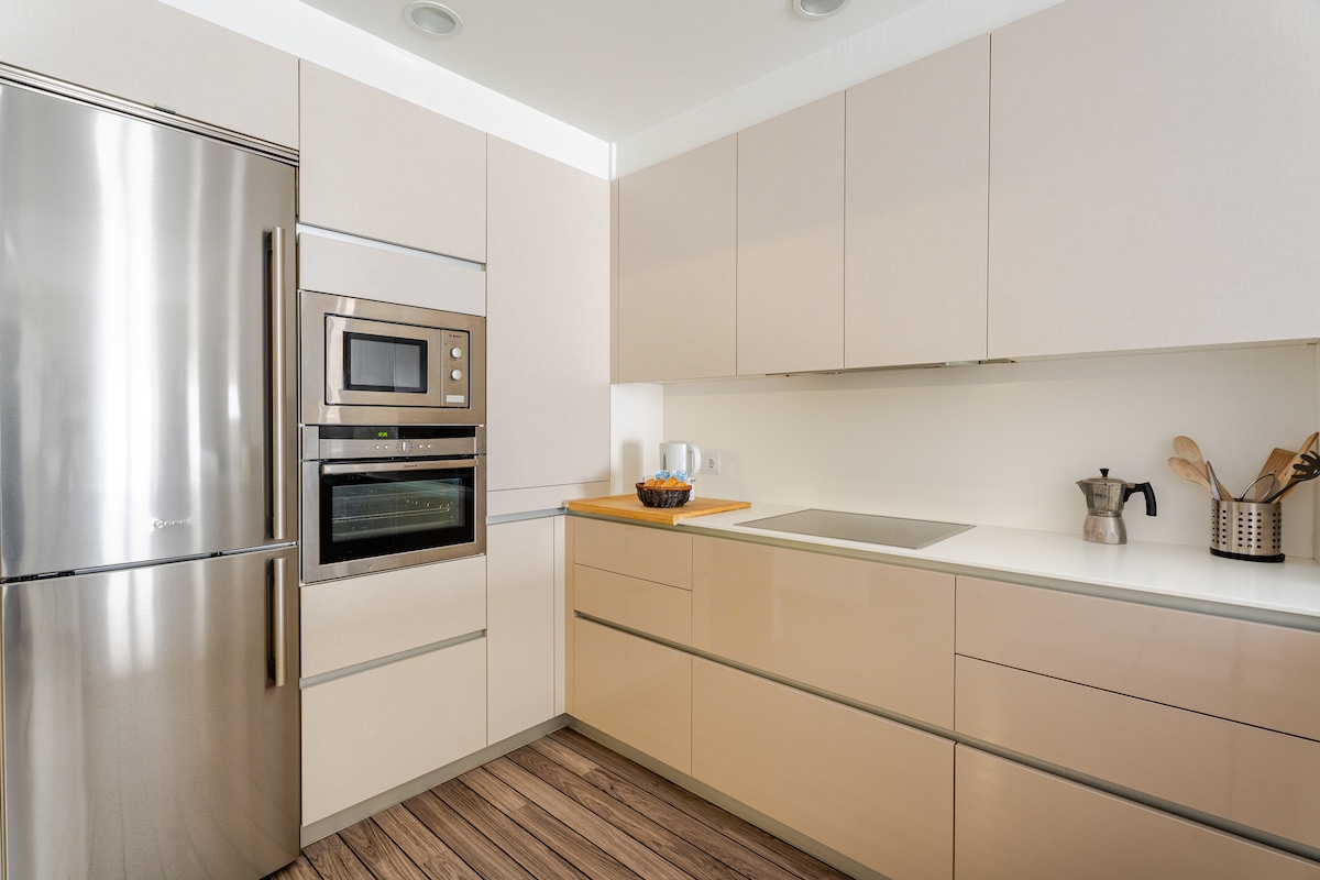 A modern kitchen features sleek cabinetry in neutral tones, paired with stainless steel appliances. A microwave and oven are built into the cabinetry, while a large refrigerator stands nearby. A wooden cutting board holds a bowl of fruit, adding a touch of warmth to the space.