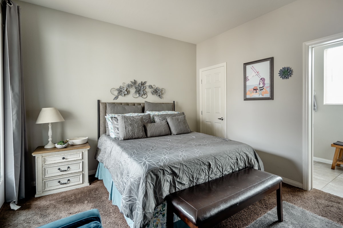 A well-appointed bedroom features a spacious bed with decorative pillows and a textured blanket. A bedside lamp and a small side table add functionality. Light-colored walls enhance the space, while a door leads to the bathroom, and an art piece is displayed on the wall.