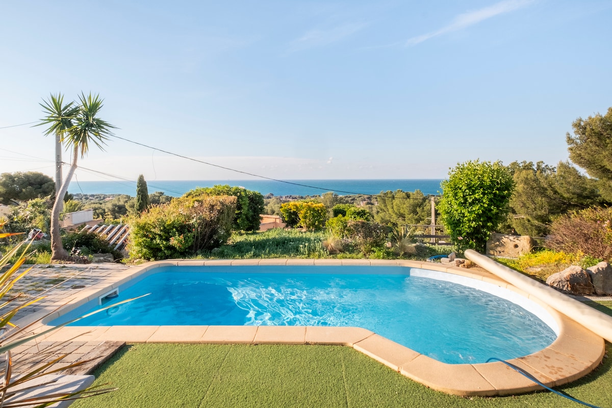 A clear view of a swimming pool is presented, bordered by lush greenery and flowers. In the background, the sea can be seen, merging with the blue sky. The setting offers a peaceful outdoor environment, ideal for relaxation.