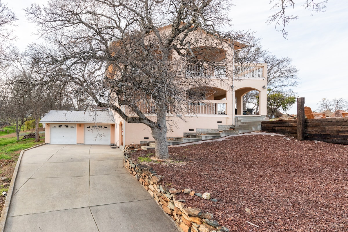 A two-story house is set on a spacious lot with gravel landscaping. The exterior is painted in a soft hue, complemented by a wrap-around balcony with railing. A driveway leads to a double garage, surrounded by mature trees and natural scenery.