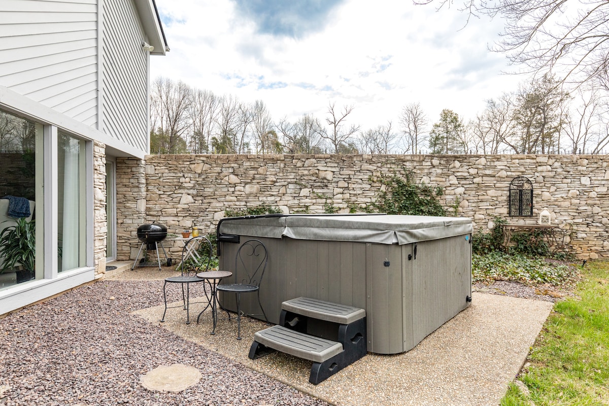 A spacious outdoor area features a large hot tub surrounded by a stone wall for privacy. Nearby, a small table and two chairs provide a cozy spot for relaxation. A grill is positioned to one side, set against a backdrop of lush greenery.