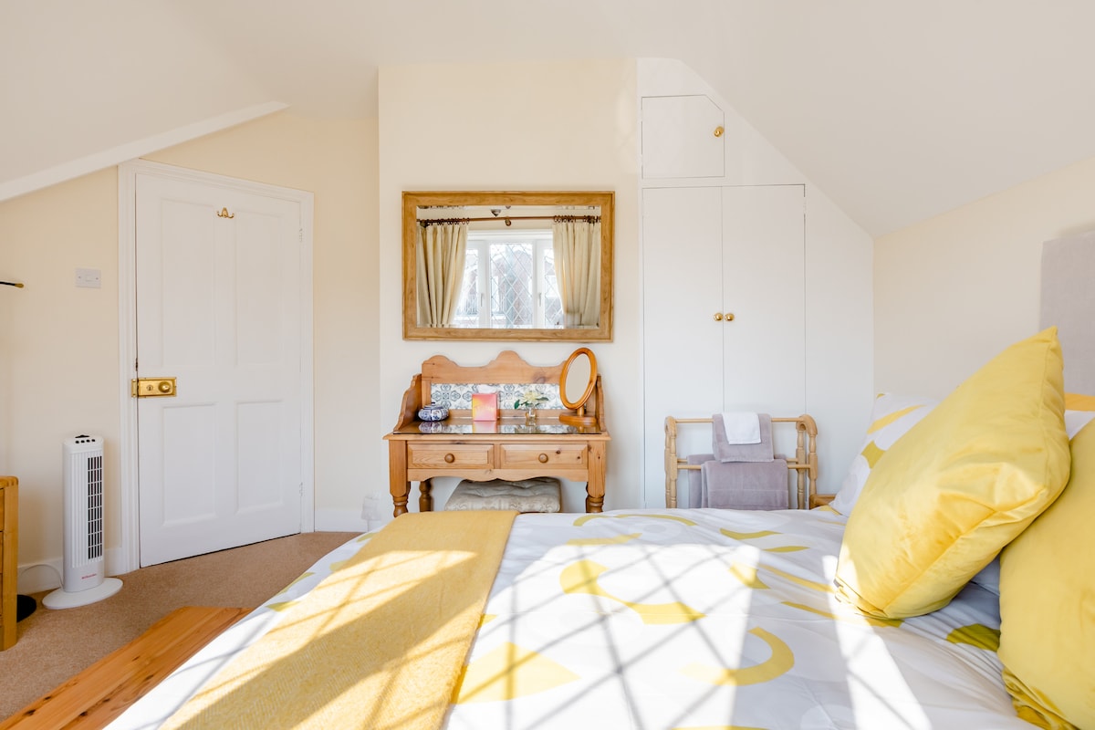 A bright and airy bedroom features a spacious bed adorned with yellow and white bedding. A wooden dresser sits against the wall with a mirror above, and a small chair is placed nearby. Morning light filters through a window covered with sheer curtains.