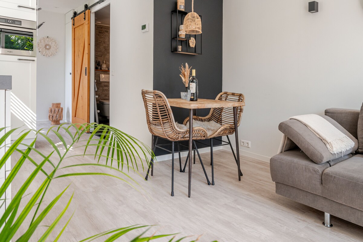 A dining area is featured with a round wooden table, accompanied by two wicker chairs. A sleek grey sofa is positioned nearby, with light streaming in from a window. Decorative elements include a wall-mounted shelf and potted plant, adding a touch of greenery to the space.