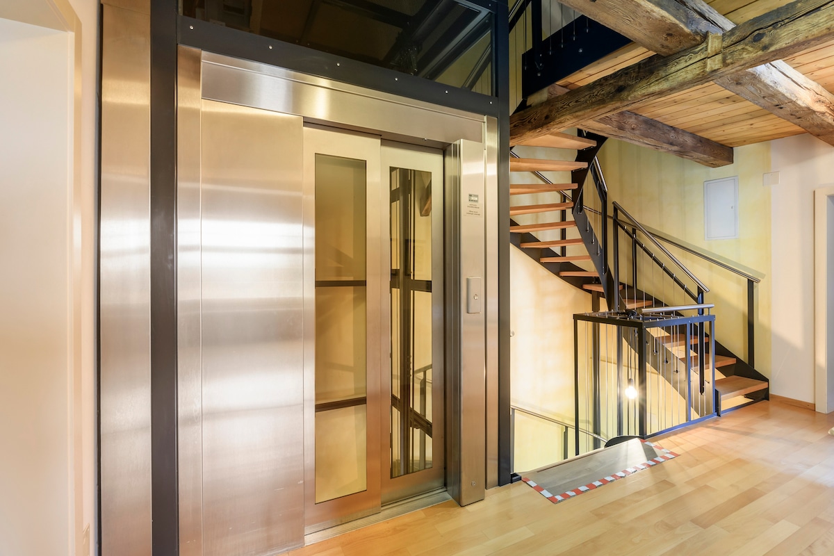 An accessible elevator is positioned against a wall with a glass door, reflecting the light from the hallway. A wooden staircase with dark railing curves nearby, leading to the upper levels. The floors feature a warm wooden finish.