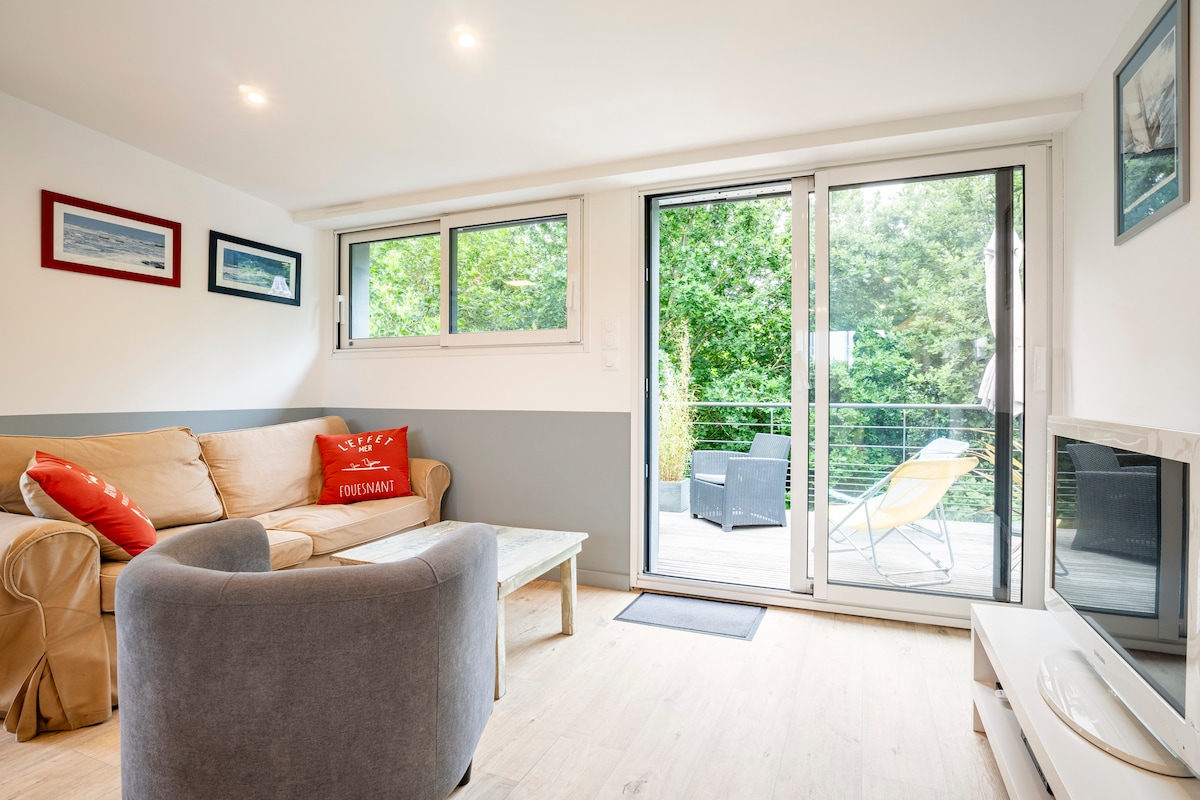A light-filled living area is presented, featuring a comfortable sofa with decorative cushions. Large glass doors provide access to a terrace, allowing natural light to illuminate the space. A neutral color palette enhances the serene ambiance, complemented by artwork on the walls.
