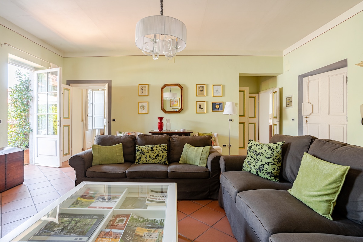 A spacious living area is furnished with a large brown sectional sofa adorned with colorful cushions. Natural light enters through double doors leading to a terrace. A glass coffee table rests on terracotta tiles, with framed art pieces visible on the walls.