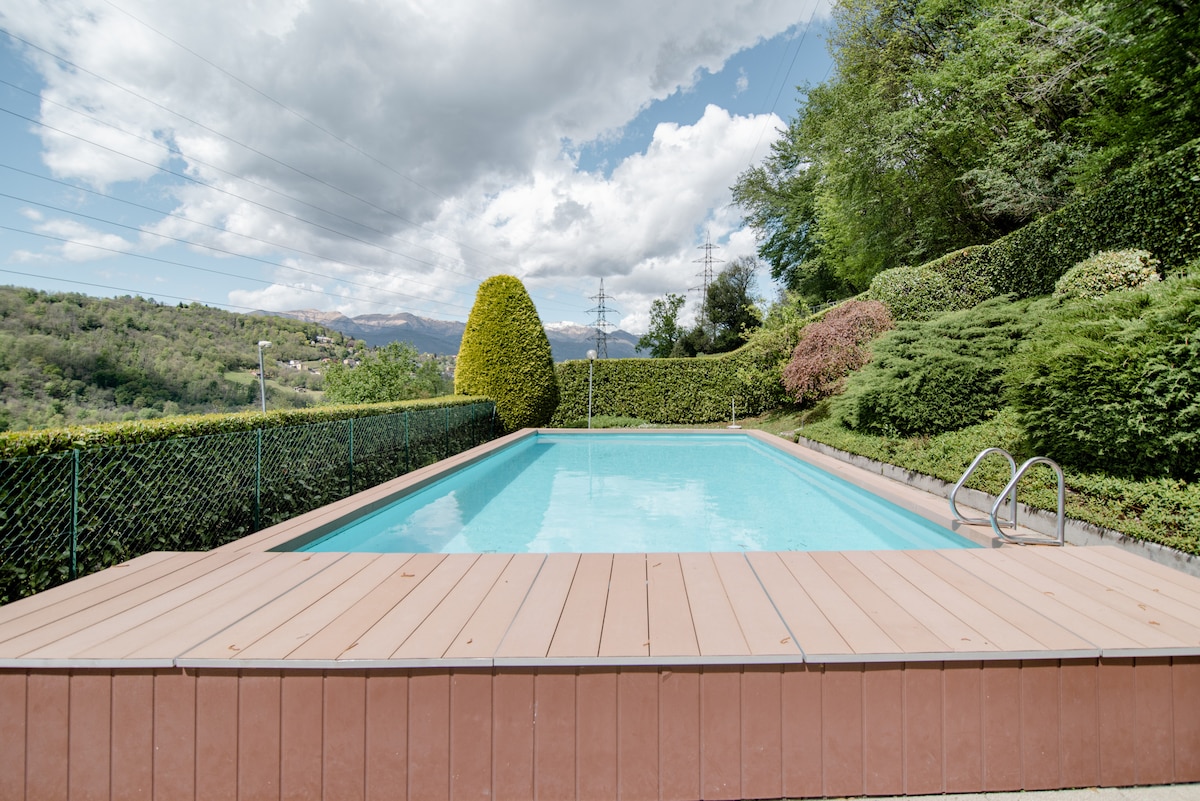 A serene outdoor pool is framed by lush greenery and hedges, with a wooden deck surrounding the water. The sky features soft clouds, casting a gentle light on the pool's clear blue surface.