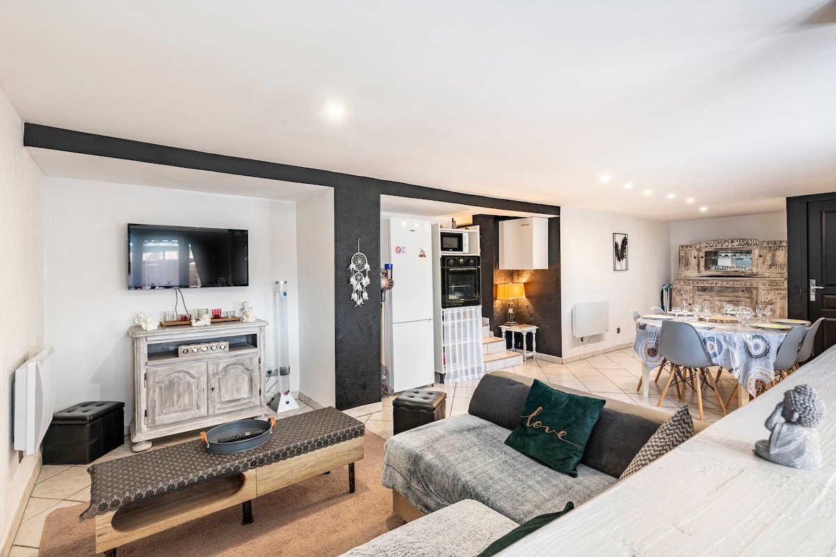 A modern living area is showcased, featuring a comfortable sofa and a coffee table. A wall-mounted television is visible, alongside a kitchen equipped with an oven and refrigerator. A dining table is situated in the corner, accommodating multiple guests. Natural light fills the space.