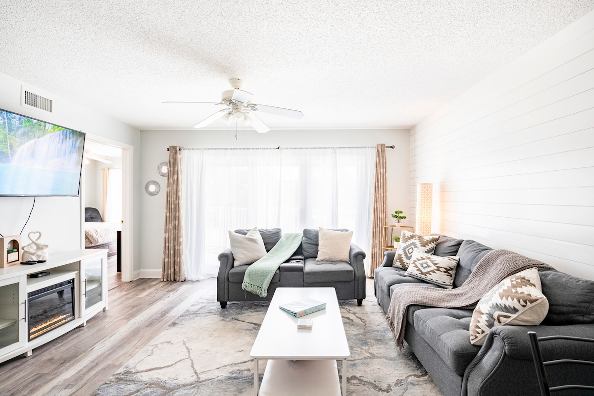 A bright living area features a light grey wall with natural lighting from large windows. Two cozy sofas, one in dark grey and the other in a mixed pattern, offer seating. A white coffee table sits in the center, complemented by a decorative rug and a fan overhead.