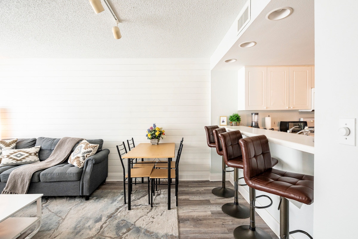 A modern living and dining area features a cozy grey sofa adorned with a blanket. A simple dining table is set beside a small flower arrangement. Barstools line the kitchen counter, providing an inviting space for meals and gatherings.