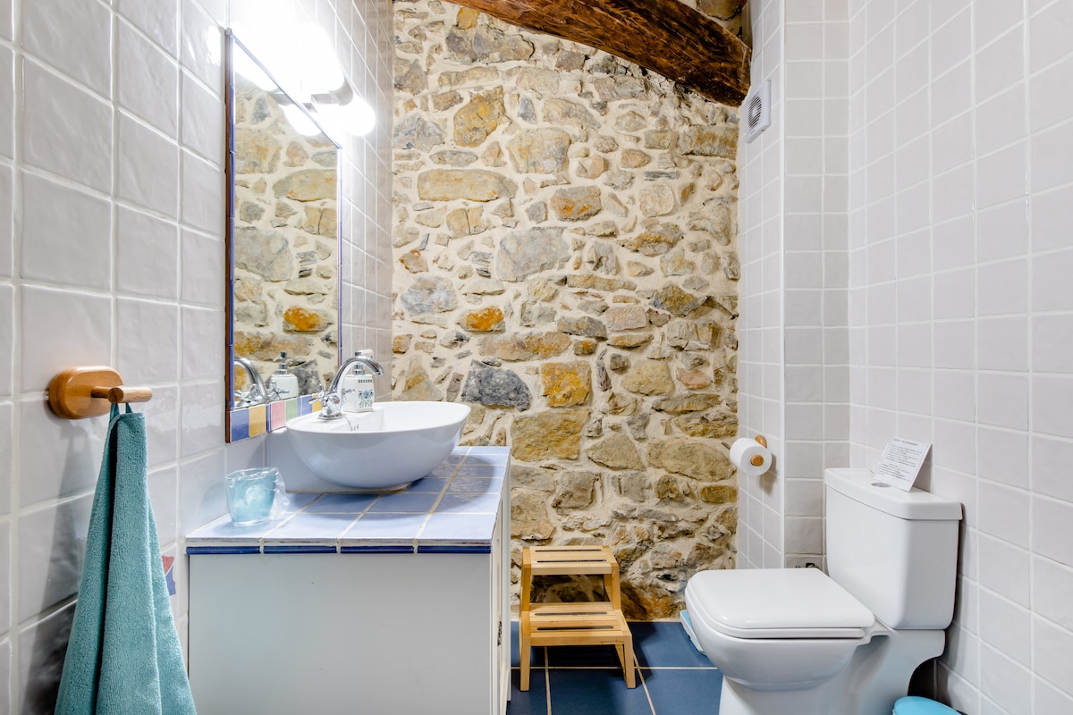 The en suite bathroom features a stone wall that adds character to the space. A modern sink rests on a tiled countertop, complemented by a large mirror above. A wooden stool is positioned nearby, and clean white fixtures enhance the bright, open atmosphere.