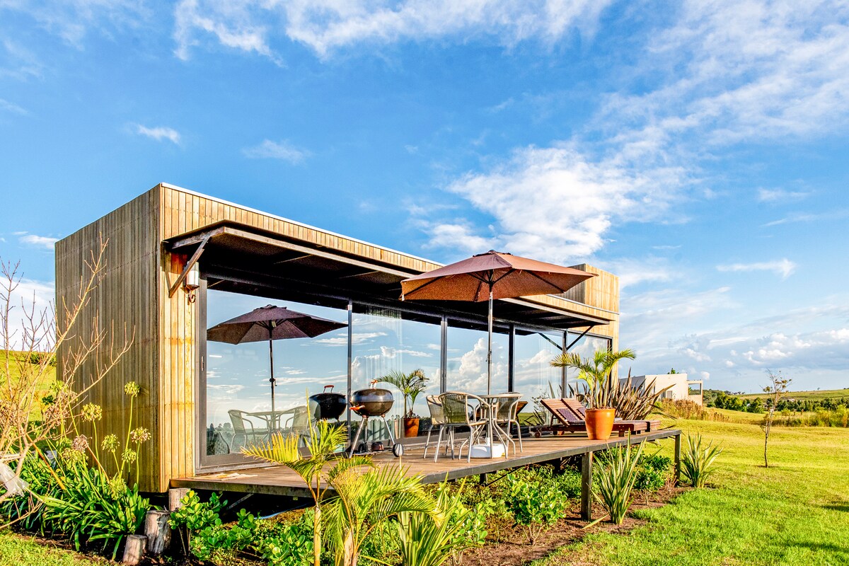 A modern container home is showcased with large glass windows and a spacious wooden deck. Outdoor seating arrangements include tables and chairs beneath umbrellas, surrounded by lush greenery. The serene landscape features an expansive sky, enhancing the connection to nature.