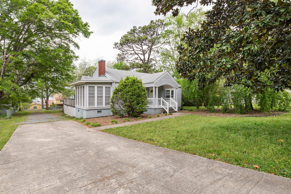A charming cottage sits surrounded by lush greenery, showcasing a front porch with steps leading to the entrance. A spacious driveway leads up to the home, providing ample parking space. Mature trees and manicured shrubs enhance the peaceful outdoor setting.