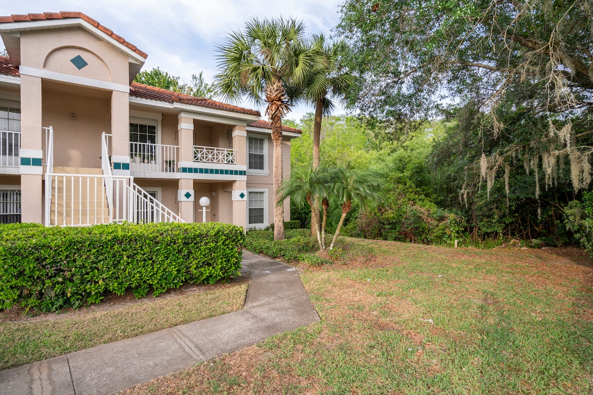 An exterior view of the apartment building showcases a two-story structure surrounded by lush greenery. Palm trees and shrubs line the walkway leading to the entrance, which features white railings and a tiled roof. The area is shaded by a canopy of trees.