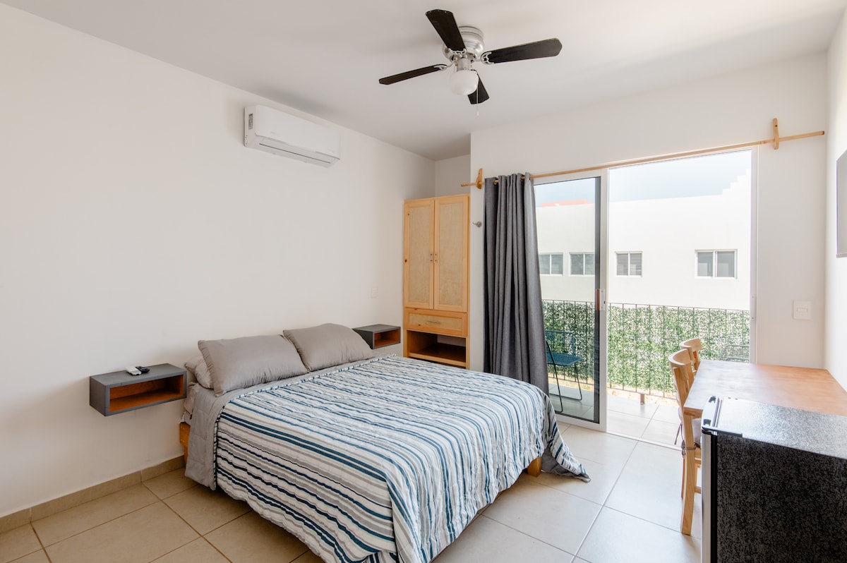 A cozy accommodation features a spacious bed with a striped blanket, accompanied by matching nightstands. A desk sits nearby, and a sliding glass door leads to a balcony. Natural light floods the room through the door, and a ceiling fan provides additional comfort.