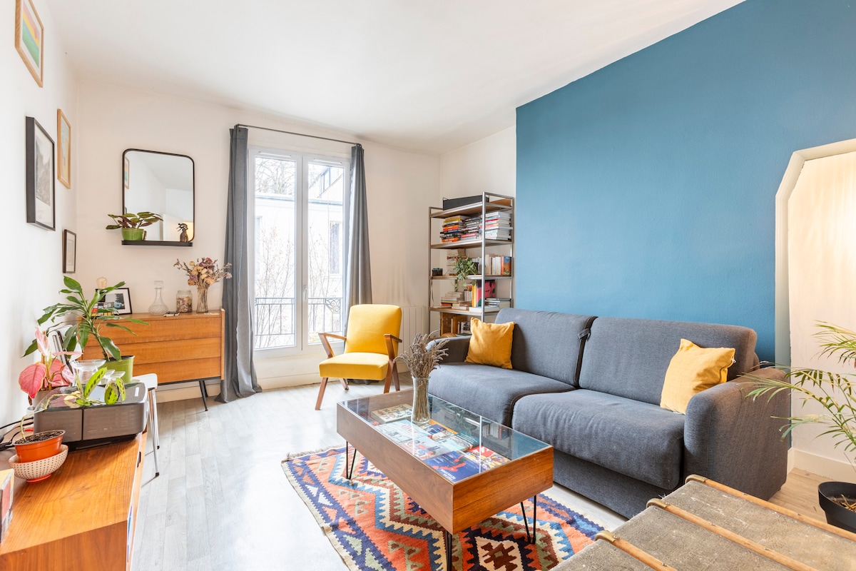 A living area is characterized by a blue accent wall and natural light pouring in through a large window. A comfortable gray sofa faces a wooden coffee table, with an inviting yellow armchair nearby. Shelves display books and decorative items, complemented by indoor plants.