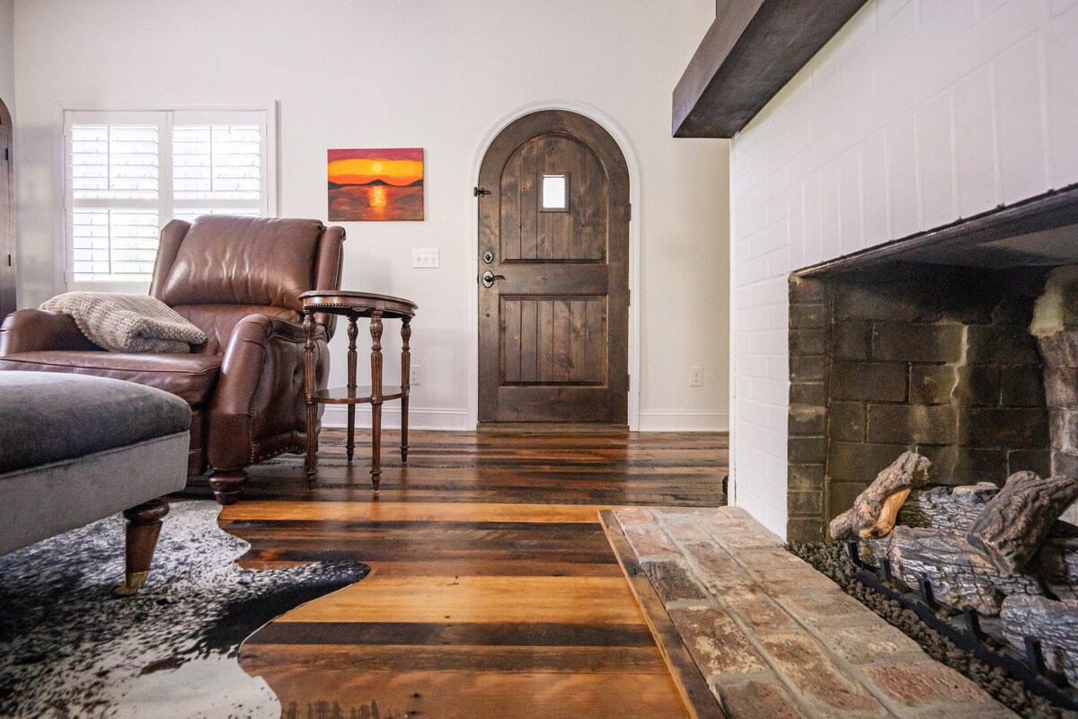 A welcoming entryway features a distinctive arched wooden door and an inviting leather recliner next to a small side table. A fireplace with exposed brick and logs complements the natural wood flooring that displays an interesting pattern.