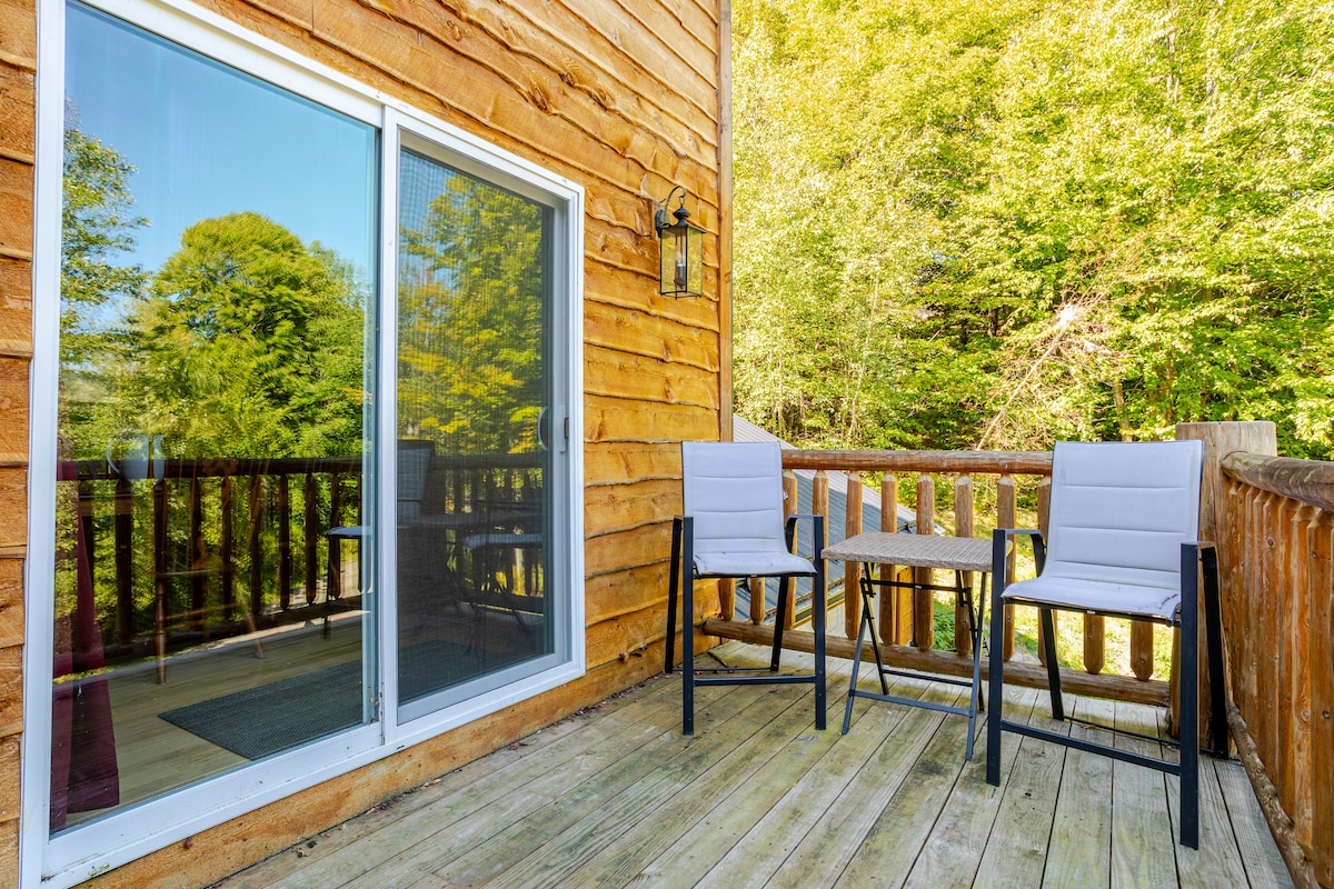 A private outdoor deck is shown, featuring two modern chairs alongside a small table. The wooden railing provides a natural view, surrounded by lush green trees and reflecting the serenity of the surrounding area.