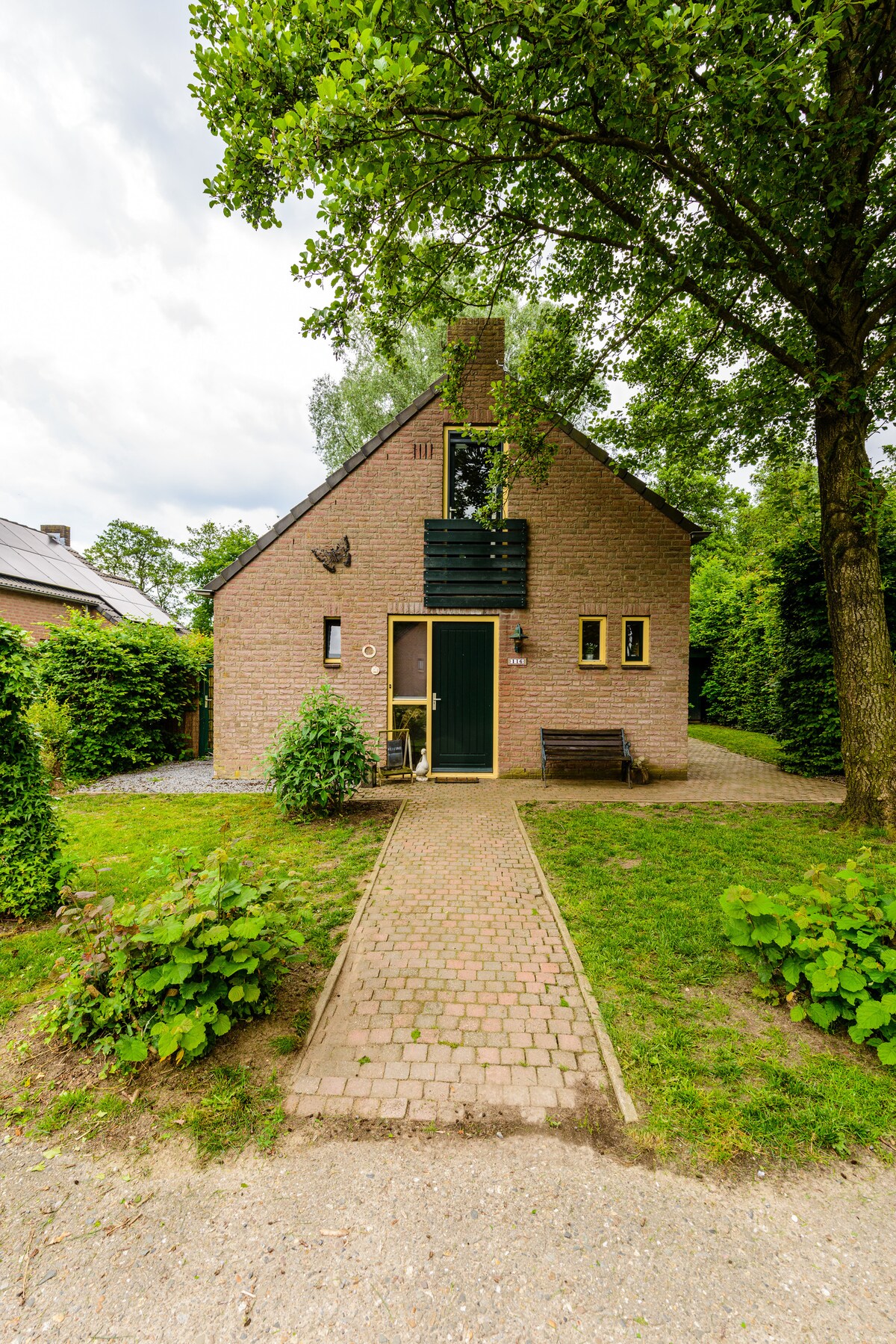 The exterior of the house features a brick facade with dark green accents and a welcoming entrance. A paved pathway leads to the front door, bordered by greenery and shrubs. The house is nestled among trees, providing a natural, tranquil setting.