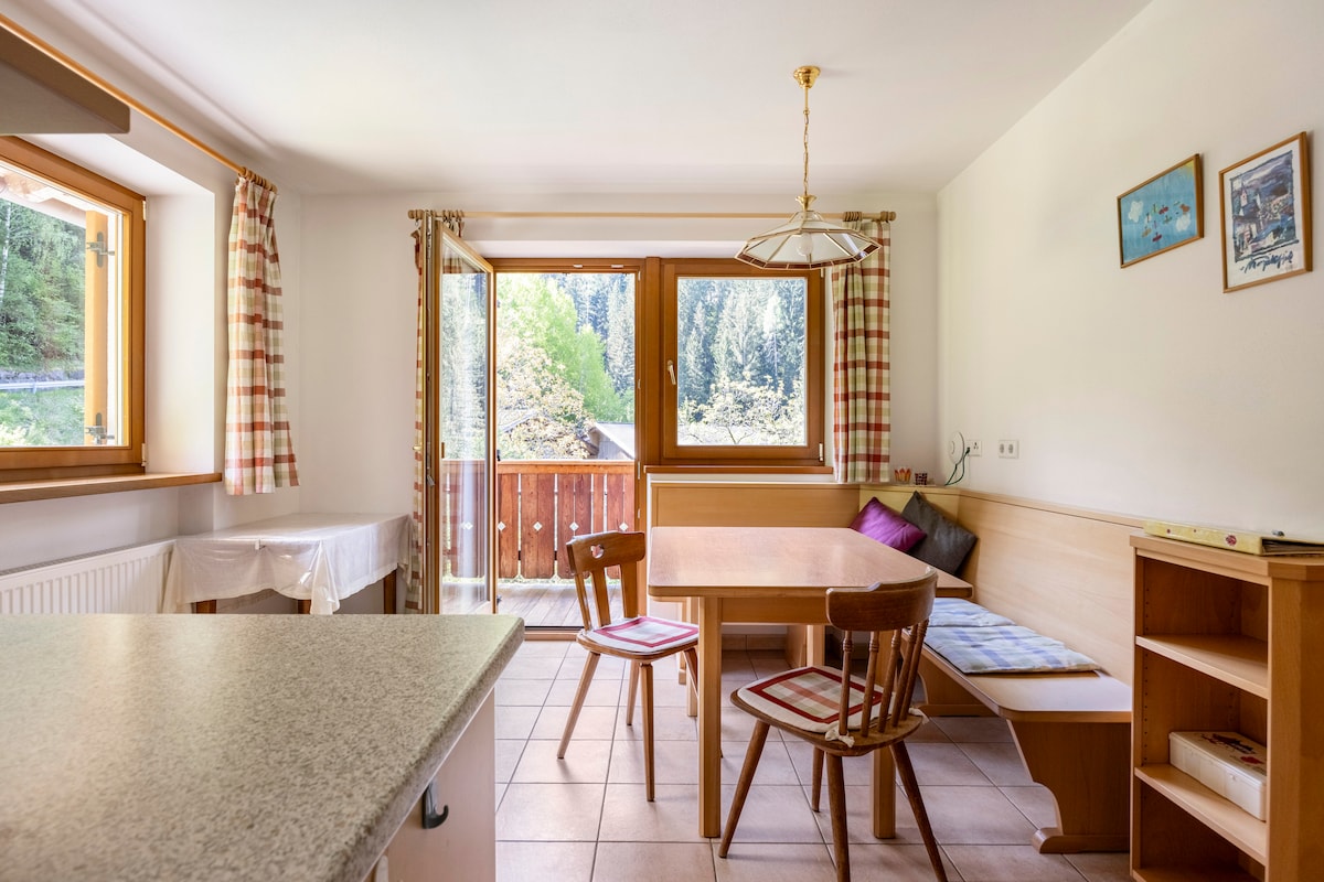 A bright dining area features a wooden table with seating for four and a bench, illuminated by natural light from large windows. A sliding door leads to a balcony with views of the surrounding nature. The space is complemented by light tile flooring and simple decor.