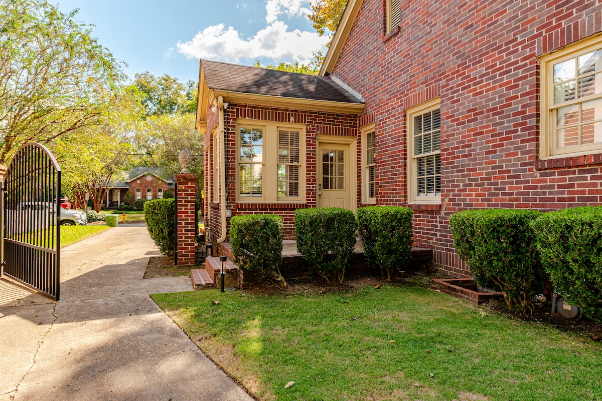 The exterior of a 1930s brick home is presented, showcasing well-maintained landscaping and a welcoming front entrance. The manicured bushes border the pathway leading to the door, while the charming brick façade reflects the character of the Historic Old Cloverdale neighborhood. A private driveway is visible.