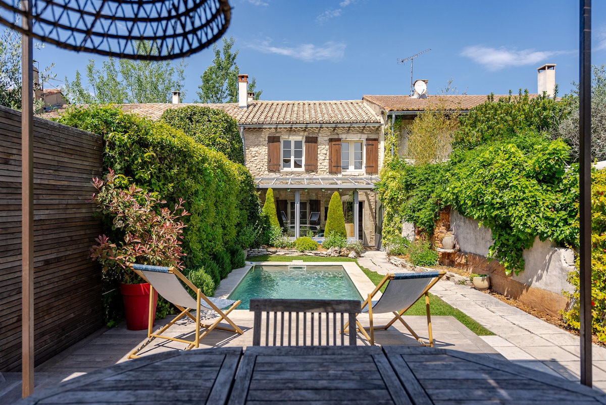 A lush garden area is visible, featuring a clear pool framed by well-maintained greenery. Two deck chairs are positioned invitingly near the water. The home façade, highlighted by warm tones, is partially obscured by vibrant foliage, creating a serene outdoor retreat.