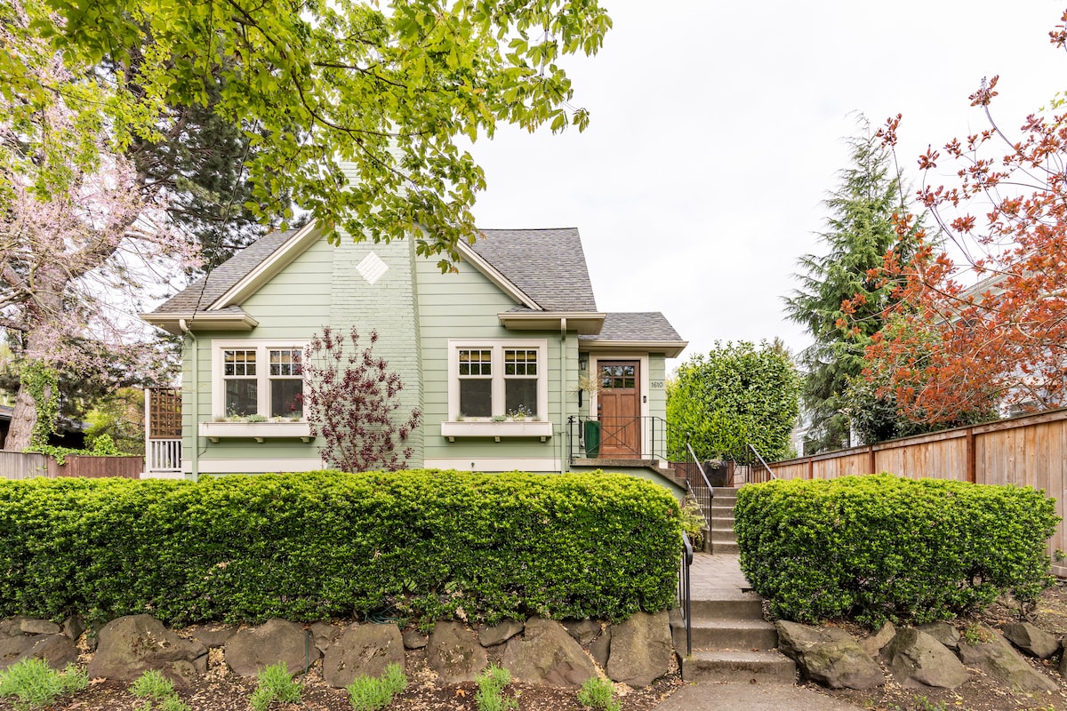The exterior of a light green craftsman-style house is framed by neatly landscaped bushes and trees. A pathway leads up to the wooden front door, with a few steps visible. The property is set back from the street, demonstrating a quiet, residential setting.