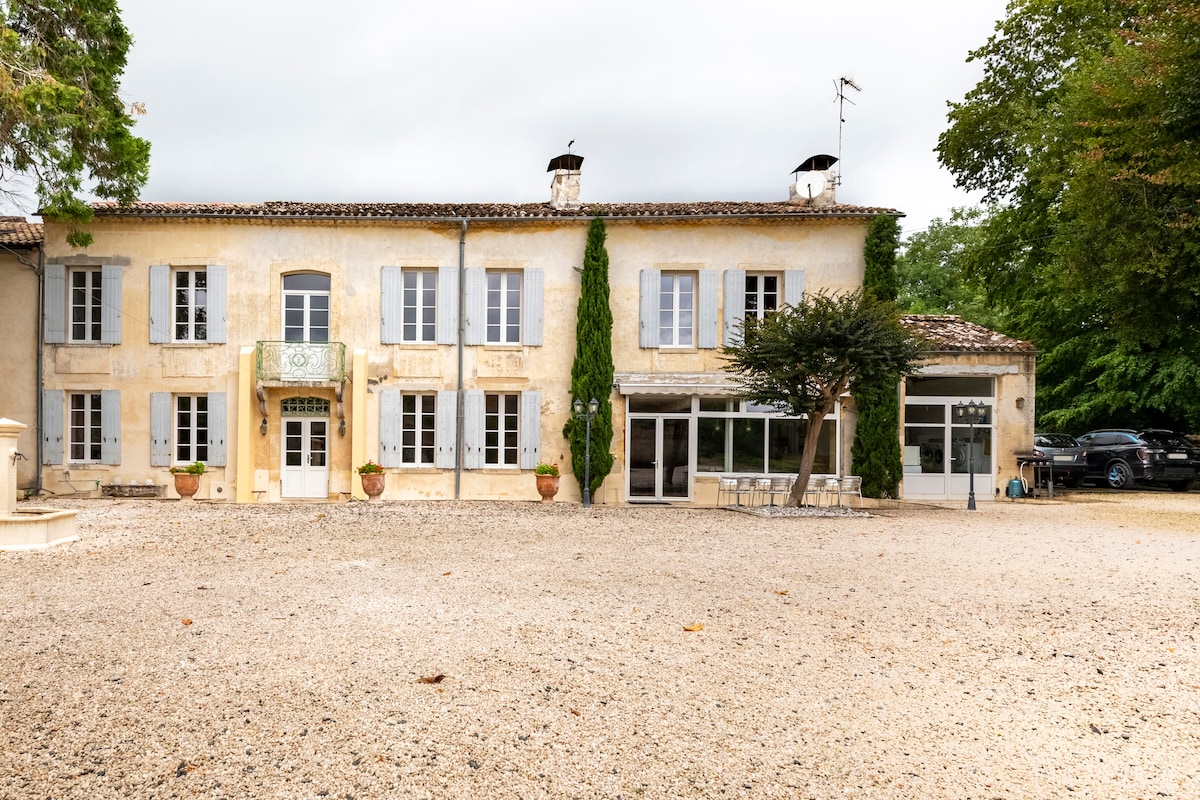 The exterior of the two-story house features soft yellow stone walls and a gravel courtyard. Large windows reflect natural light, while potted plants are positioned near the entrance. A shaded patio area is visible to the right, accompanied by greenery and trees.