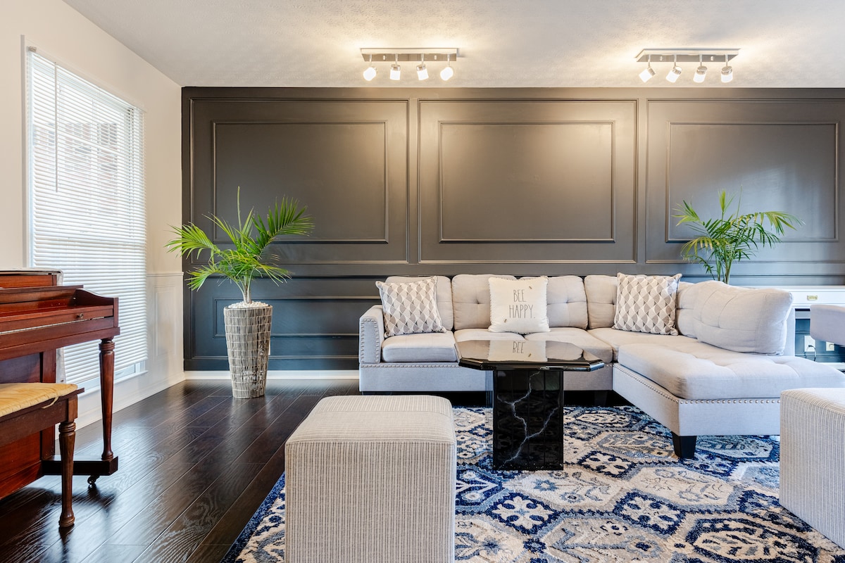 A bright living area features a light-colored sectional sofa and a coffee table atop an intricate area rug. Decorative plants are positioned beside the seating, and a wooden piano is present, adding to the room's character.