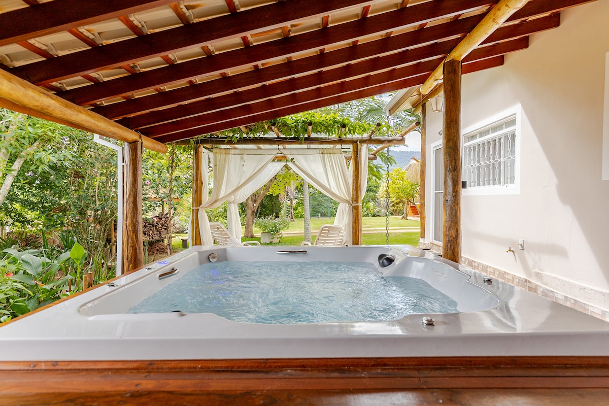 A relaxing spa area is framed by a wooden structure, featuring a jetted hot tub. Two light-colored lounge chairs are positioned beside the tub, with a sheer curtain providing partial privacy. Lush greenery surrounds the space, enhancing the natural ambiance.