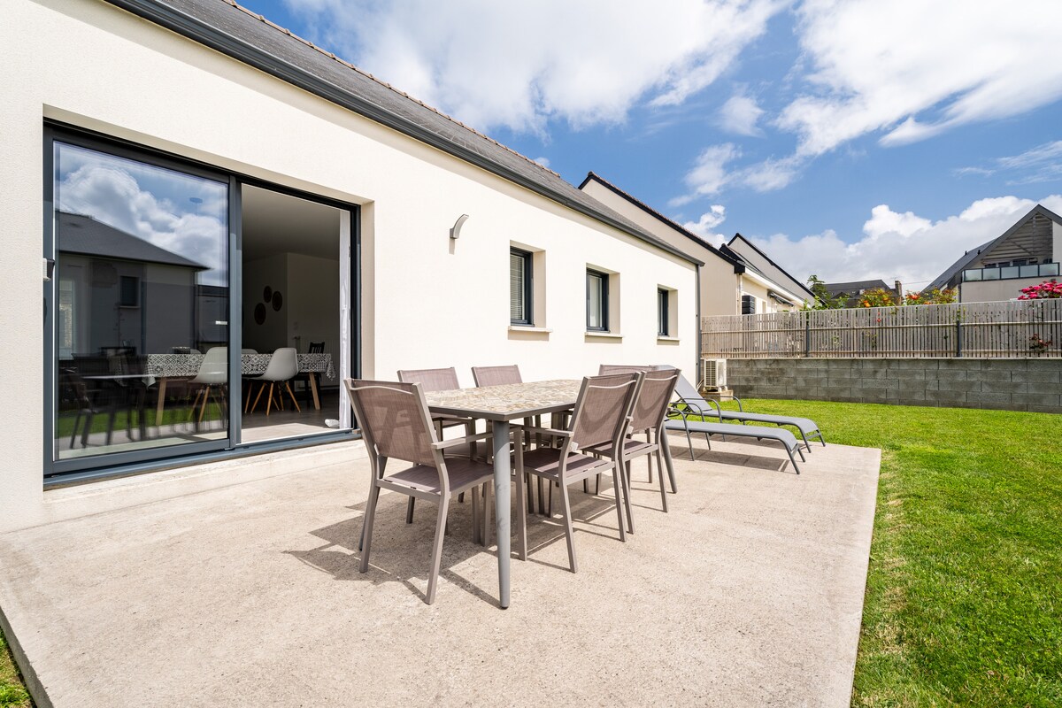 A spacious terrace is visible, featuring a large table surrounded by six chairs. Two sun loungers are positioned nearby on the well-maintained lawn. The terrace offers direct access to the bright interior through large glass doors.