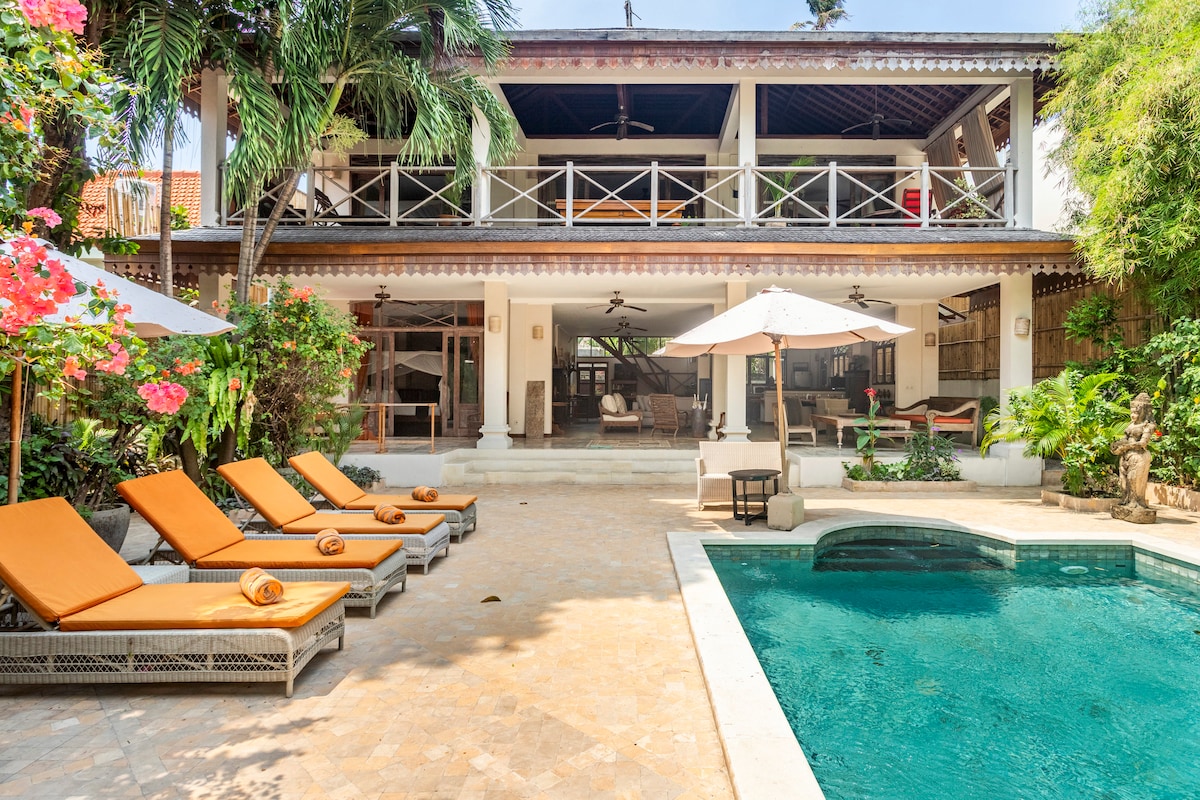 A serene outdoor area features a private swimming pool surrounded by tropical plants and lounge chairs with orange cushions. The two-story villa is visible in the background, showcasing a spacious patio with ceiling fans and ample seating options.