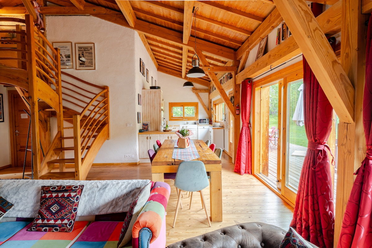 A spacious open-plan living area showcases wooden beams and a high ceiling, enhancing the bright atmosphere. A large dining table with colorful chairs is centered in the room, surrounded by large windows that open to the garden. A cozy sofa is adorned with vibrant cushions.