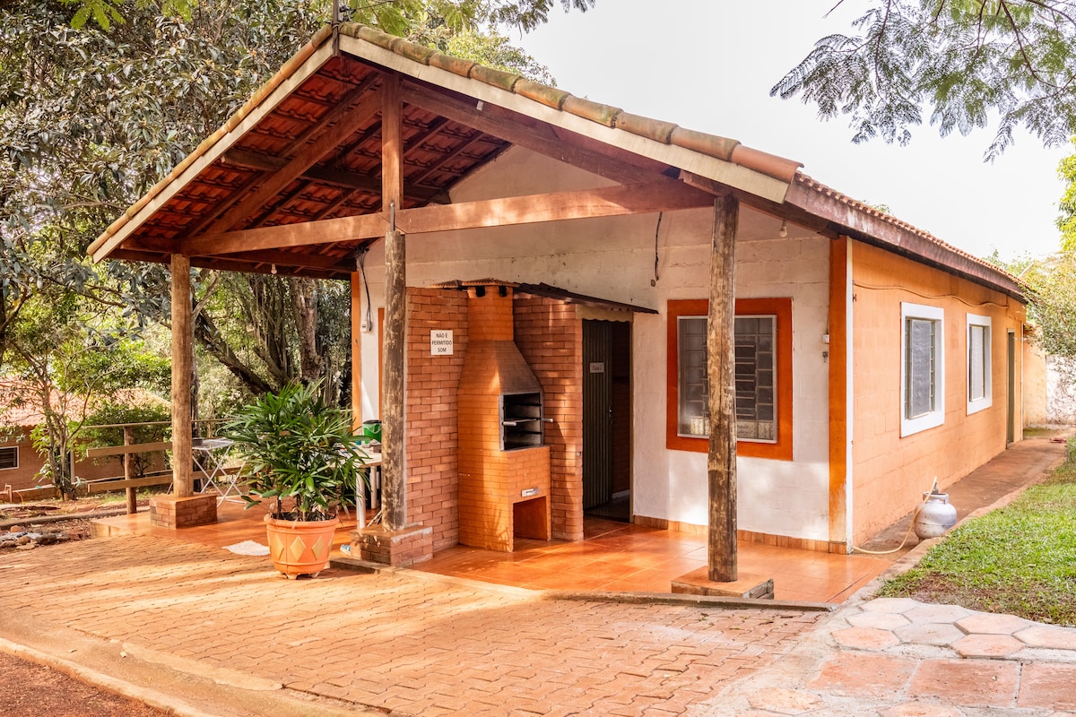 The exterior of a single-story house is shown, featuring a covered porch supported by wooden beams. A brick barbecue area is present, along with plants placed in decorative pots. Large windows allow natural light to enter, surrounded by greenery in a tranquil setting.